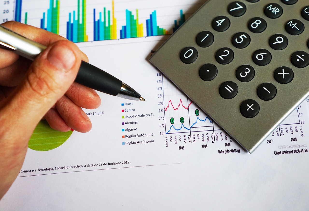 A hand holding a pen over printed bar and line charts beside a calculator